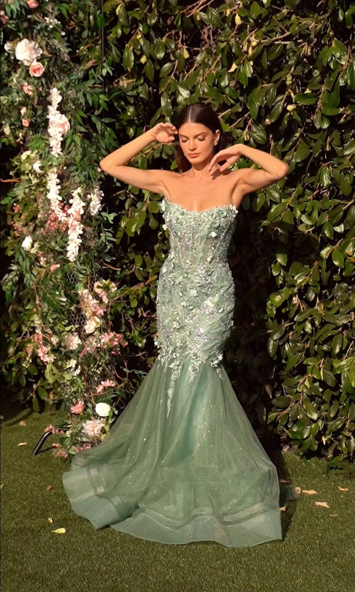 A woman in the Long Sleeve Long Formal Dress: Andrea & Leo MA101 stands before greenery and flowers, adjusting her hair.